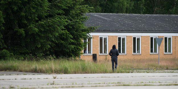 Ein Mann mit Kapuzenpullover vor einem flachen, barackenartigen Gebäude, links Gebüsch, davor eine Betonfläche, auf der Gras wuchert