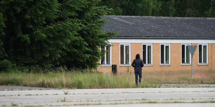 Ein Mann mit Kapuzenpullover vor einem flachen, barackenartigen Gebäude, links Gebüsch, davor eine Betonfläche, auf der Gras wuchert