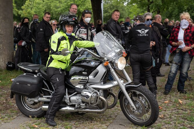 Marie-Luise Strack-Zimmermann sitzt auf einem Motorrad, sie fährt gleich los. Im Hintergrund steht eine Gruppe Männer in Biker-Kleidung