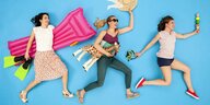 Drei Junge Frauen rennen vor einem blauen Hintergrund und tragen Assecooires für enen Strandaufenthalt