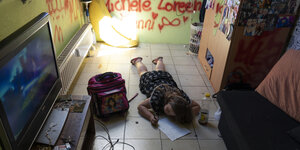 Ein Kind liegt bäuchlings auf dem Boden, links ein TV, im Hintergrund Graffiti an der Wand
