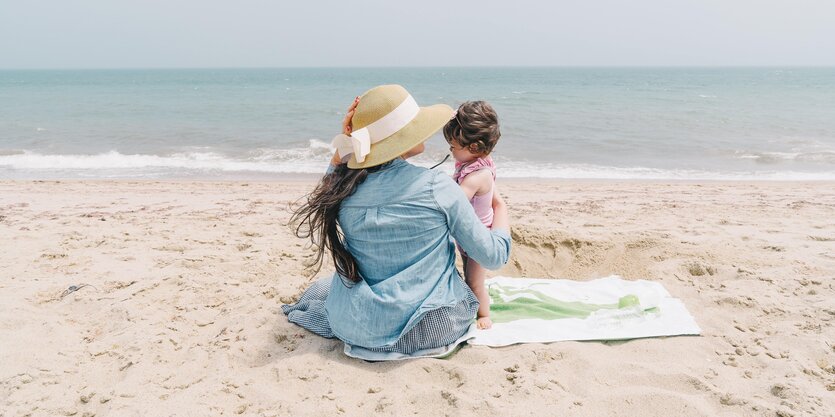 Eine Frau in blauem Kleid mit Strohhut und ein Kind zusammen an einem Sandstrand am Meer