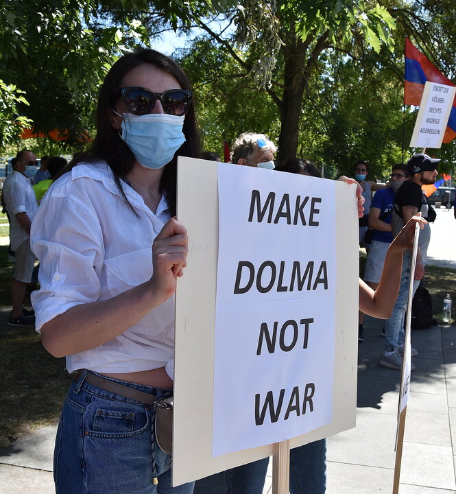 Eine junge Frau demonstriert gegen die Kämpfe am Berg Karabach zwischen Armenien und Aserbaidschan. Sie trägt einen Mundschutz und hält ein Schild vor sich auf dem steht: „Make Dolma not war“.