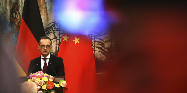Heiko Maas vor den Flaggen Chinas und Deutschlands, dahinter ein Wandgemälde, das einen Wald zeigt.