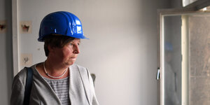 Katrin Lompscher blickt aus dem Fenster eines Rohbaus. Sie trägt einen blauen BauarbeiterInnenhelm