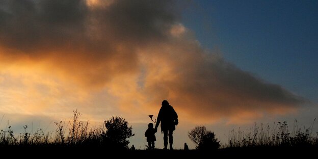 Kind läuft mit Elternteil auf einer Wiese in den Sonnenuntergang