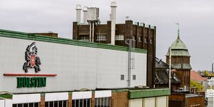 links weiße Schuhschachtelhalle mit grünem Rand am Dach, rechtsdahinter Backsteingebäude wie eine Burg