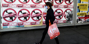 eine Frau geht mit Gesichtsmaske und Einkaufsbeutel an Protestplakaten vorbei
