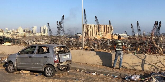 Ein zerstörtes Auto und das zerstörte Hafengelände von Beirut, im Vordergrund ein Mann, der mit dem Rücken zur Kamera steht.