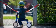Ein Kind fährt auf einem Dreirad an einem gesperrten Spielplatz vorbei.
