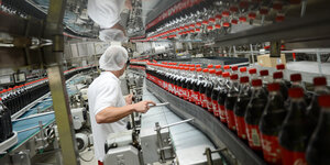 Förderband mit Coca-Cola-Flaschen in einer Fabrik, daneben ein Arbeiter