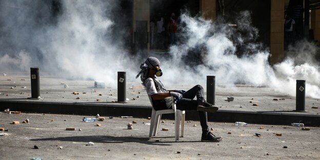 Junger Mann mit Gasmaske sitzt inmitten von Tränengaswolken auf einem Stuhl in Beiruts Straßen