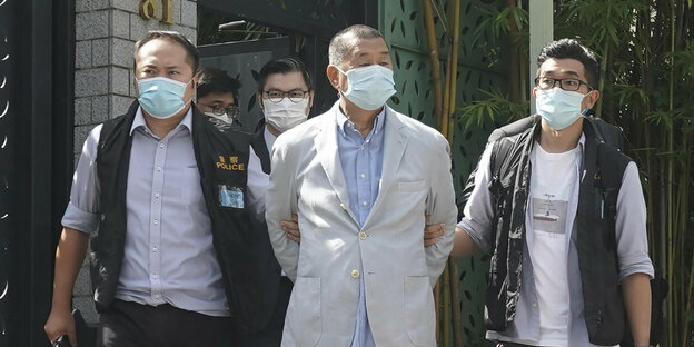 Medienunternehmer Jimmy Lai wird von zwei Polizisten abgeführt. Alle tragen Masken. Lai hat einen Anzug an, die Polizisten tragen T-Shirt oder Hemd und darüber eine Weste.