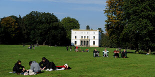 Menschen liegen auf einer Wiese, im Hintergrund ein weißes Landhaus und hohe Bäume