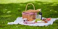 Ein Picknickkorb steht auf einer weißen Decke auf einer grünen Wiese, daneben unter anderem eine Flasche Wein und ein Stück Wassermelone