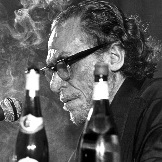 Charles Bukowski rauchend und trinkend bei einer Lesung