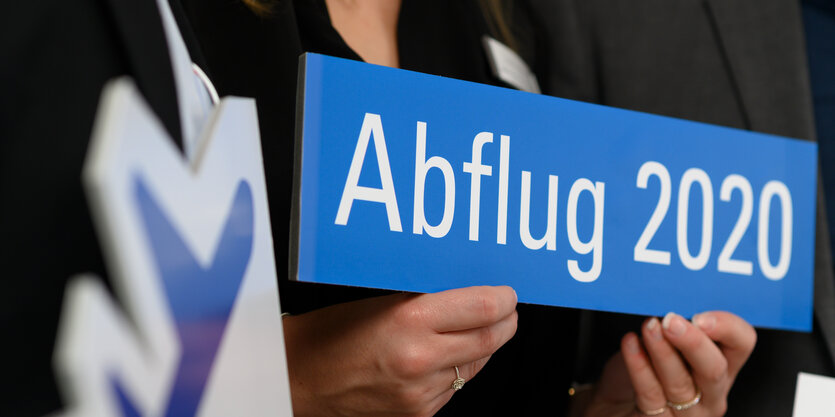 zwei Hände halten ein Schild mit der Aufschrift "Abflug 2020", links davon ist ein Teil des Flughafen-Logos zu sehen