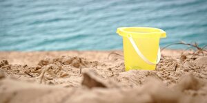 Ein gelber Eimer im Sand, im Hintergrund Wasser