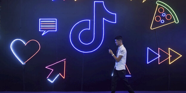Ein Mann geht an einer Wand entlang, auf der Neonsymbole die App TikTok bewerben