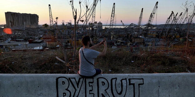 Ein Mann sitzt fotografierend auf einer Mauer vor zerstörten Kränen