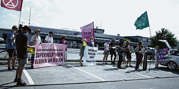 Demonstranten mit Fahnen, Plakaten und Musikinstrumenten stehen vor dem Flughafen Lübeck.