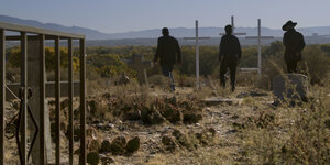 Drei Männer sind in einer kargen Landschaft zu sehen. Man sieht nur ihren Rücken. sie laufen über eine Friedhof, im Hitnergrund sind Berge und blauer Himmel