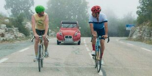 Zwei Männer auf rennrädern auf einer Alpenstraße, im Hintergrund fährt ein roter 2CV