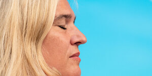 Melanie Huml im Profil mit schmerzlich geschlossenen Augen, hinter ihr ein greller, hellblauer Hintergrund
