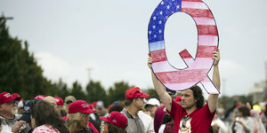 Ein Mann hält ein Q in die Luft, das in den Farben der US-Flagge ausgemalt ist. Um ihn herum viele Menschen mit Make-America-Great-Again-Kappen.