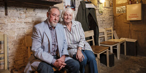 Ein Mann und eine Frau sitzen eng nebeneinander auf Stühlen in einem Keller