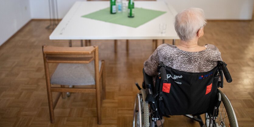 Eine alte Frau sitzt im Rollstuhl allein an einem Tisch