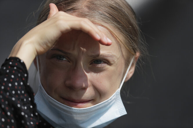 Greta Thunberg stllt die Hand schützend vor's Gesicht, um nicht von der Sonne geblendet zu werden. Ihre Atemschutzmaske ist heruntergezogen