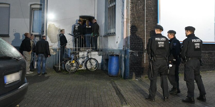 Polizisten in Uniform und weitere Männer stehen nachts vor einem Haus. Einige von ihnen sind gerade im Begriff, hineinzugehen.