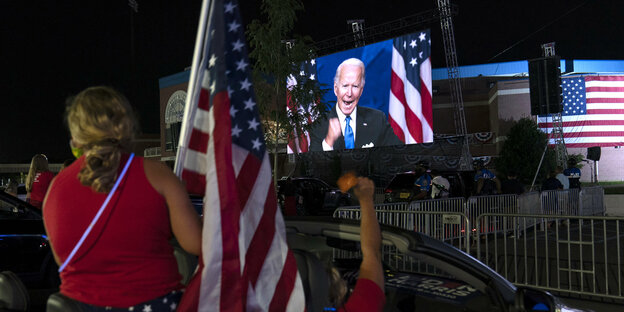 Menschen mit USA-Flaggen vor einem TV-Bildschirm, der Joe Biden zeigt.