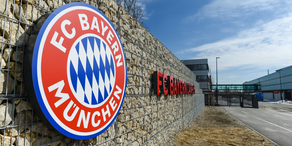 Eine Steinmauer mit Bayern-Logo und Schriftzug, rechts eine kleine Straße und Gebäude