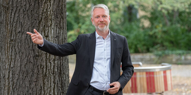 Der neue Berliner Bausenator Sebastian hat Scheel hat kurze graue Haare und einen kurzen Bart. Er trägt einen dunklen Anzug und ein hellblaues Hemd und ist schlank. Er steht vor einem Baum und hat die rechte Hand zur Seite erhoben.