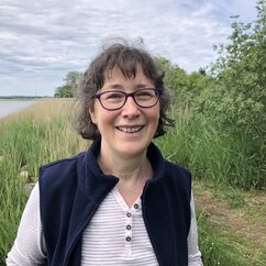 Ein Portrait von Annegret Erb vor einem Gewässer in Kiel. Sie lächelt, trägt eine Brille, kinnlange braune Haare und einen Pony