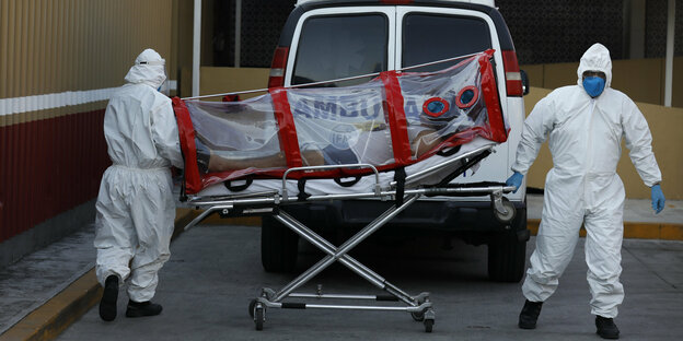 Zwei Mediziner im Ganzkörper-Schutzanzug und mit Maske schieben einen Corona-Patienten auf einer Liege zum Fahrzeug