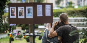 Ein Paar sitzt auf einer Bank vor einem Aufsteller mit Plakaten der Opfer von Hanau