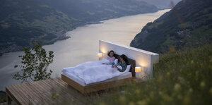 An eienm Berghang steht ein Doppelbett auf einer Terrasse aus Holzbohlen. In dem Bett liegen ein Mann und eine Frau.