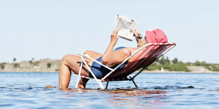 Frau liest Zeitung im Liegestuhl im Wasser.