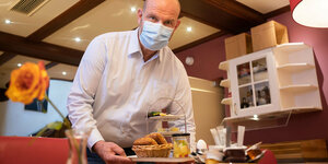Ein Mann mit Mundschutzmaske trägt ein Tablett mit einem Frühstück