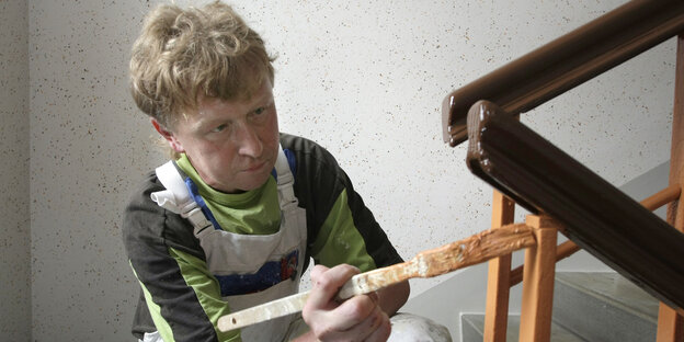 Ein Mann kniet und streicht dabei ein Treppengeländer mit hellbrauner Farbe in einem Innenraum