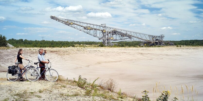 ein Fahrradfahrer und eine Fahrradfahrerin stehen vor einer Abraumförderbrücke in der Lausitz