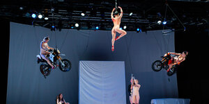 Auf einer Bühne mit ungekleideten Performerinnen schweben einige in der Luft, auf Motorrädern oder an einem Zopf.