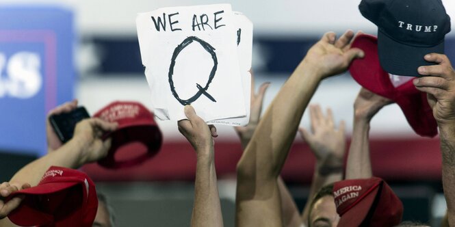 Menschen halten Mützen und ein Schild mit einem aufgemalten Q hoch.