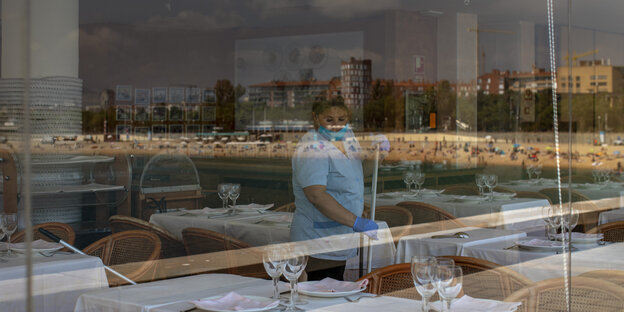Strand spiegelt sich in Fenster hinter dem eine Reinigungskraft zwischen Restauranttischen mit dem Schrubber unterwegs ist