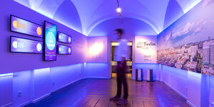 Das Bild zeigt einen in lilafarbenes Licht getauchten Raum der Ausstellung "Chaos und Aufbruch. Berlin 1920/2020" im Märkischen Museum. An der rechten Wand hängt eine große Luftaufnahme der Berliner Innenstadt Ost. Ein Mann betrachtet Bildschirme auf der