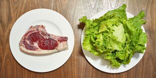 Zwei Teller stehen nebeneinander. Auf dem einen liegt ein rohes Steak, auf dem anderen ein Kopf Salat