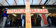 Ein Holzkreuz mit dem Namen und der Todestag. und -ort von Oury Jalloh. Dahinter Protestierende unter dem Vordach des Bahnhofs Dessau